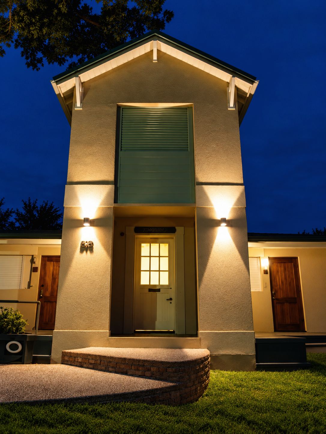 Entrada de un bungalow dúplex de noche, con las luces de la fachada encendidas y la ventana de la puerta iluminada